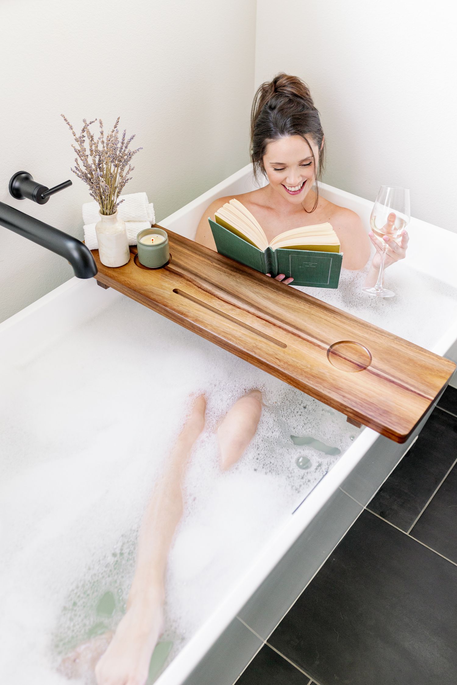 Person enjoying relaxing bath with wooden caddy tray holding book, glass of wine, and decorative vase with dried flowers.