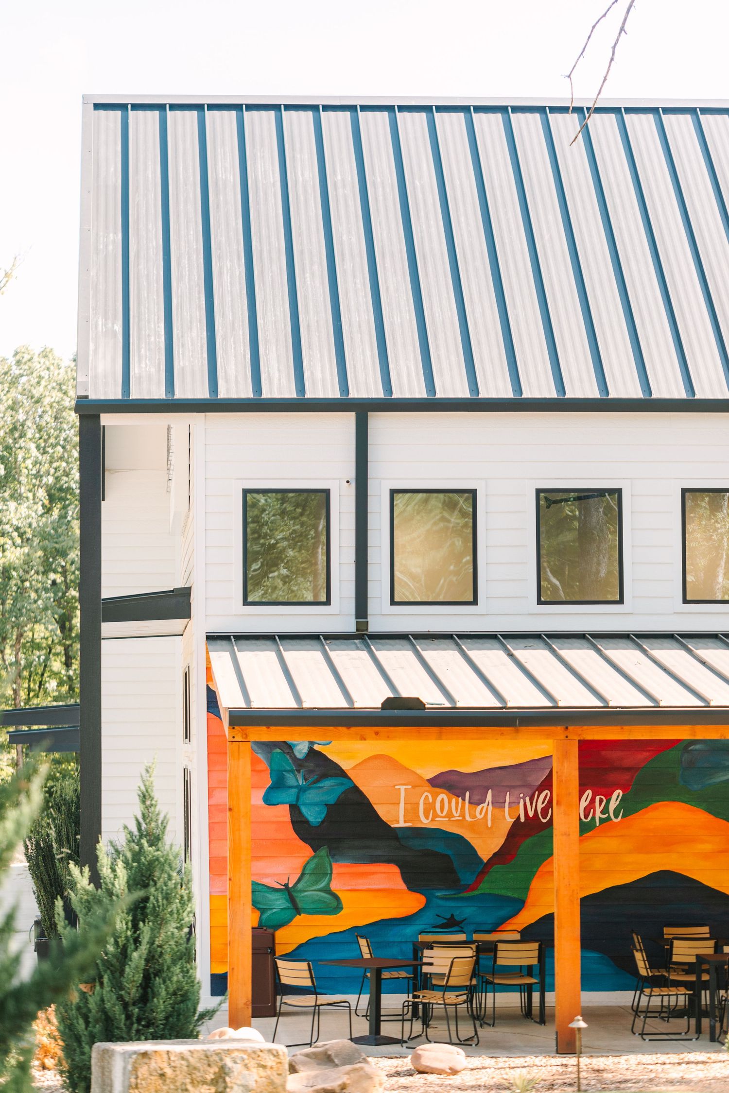A modern white building with a blue metal roof features a vibrant orange mural wall and outdoor cafe seating area.