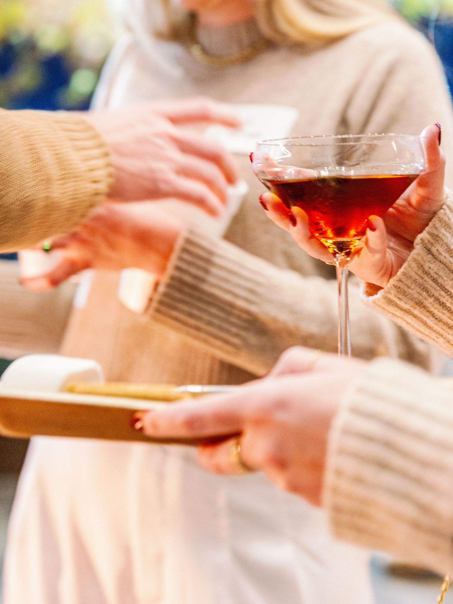 Hands holding a glass of red martini and wearing cozy beige sweaters during a social gathering.