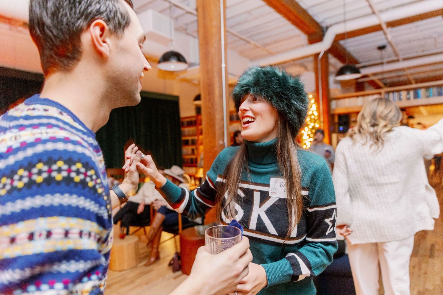 Two people in winter sweaters smile and laugh while dancing together at an indoor social gathering.