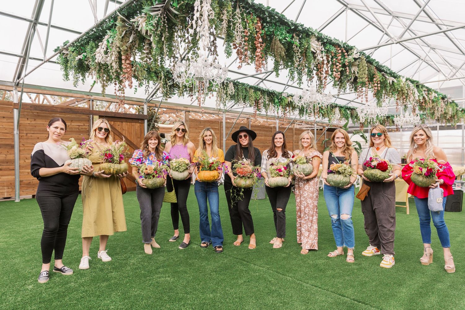 A group poses holding floral bouquets inside a greenhouse with hanging greenery and chandelier lighting above.