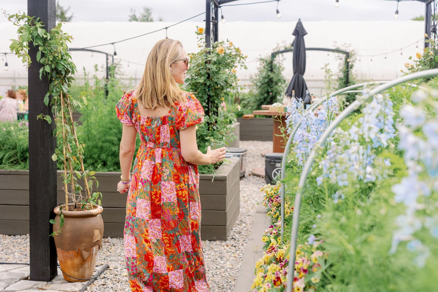 A red floral maxi dress stands out in a lush garden patio with potted plants and climbing vines.