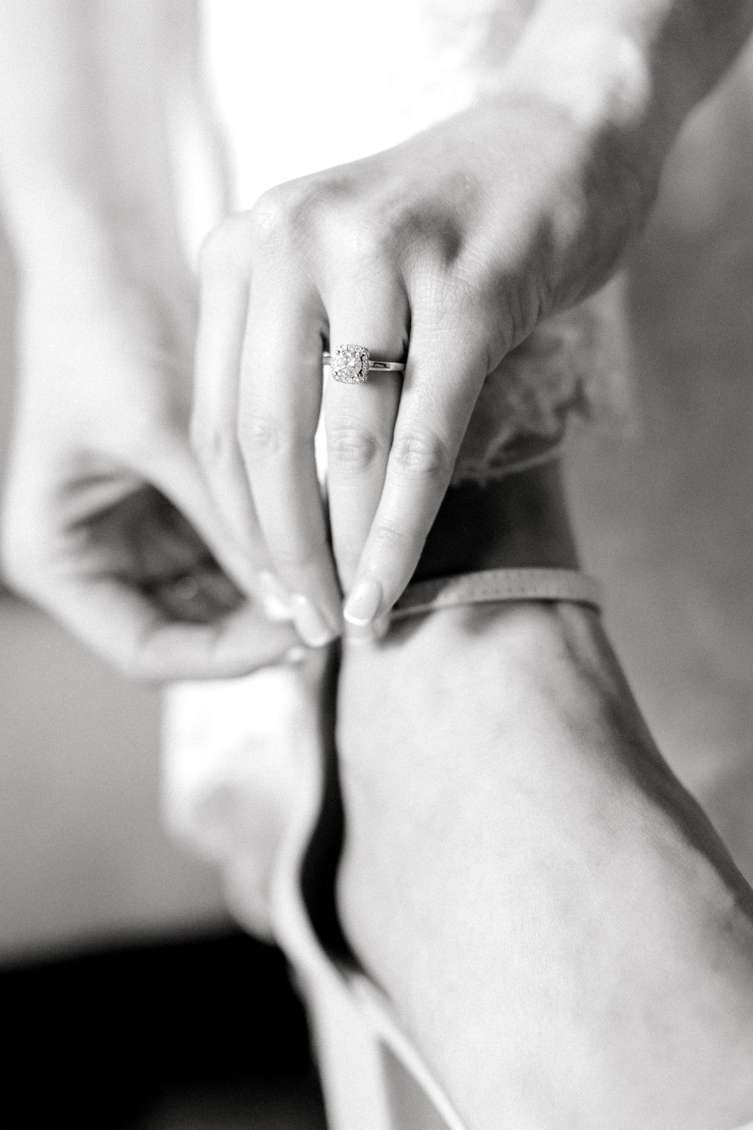 A close-up black and white photo of hands fastening a ballet shoe strap, with an engagement ring visible.