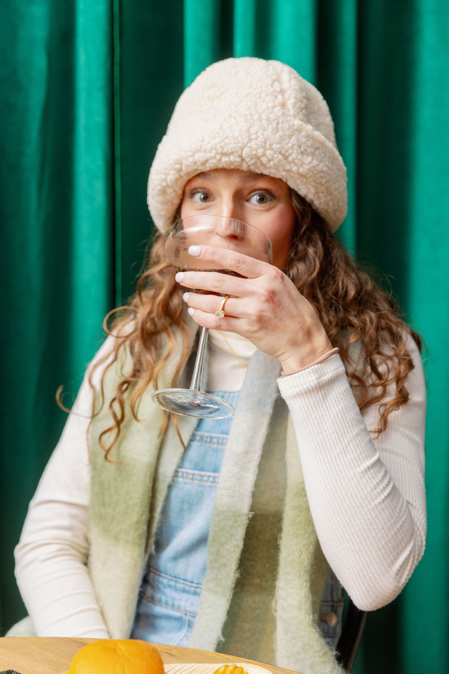 Person in winter attire with curly hair wearing a cream beanie and denim overalls sips from a drink against green backdrop.