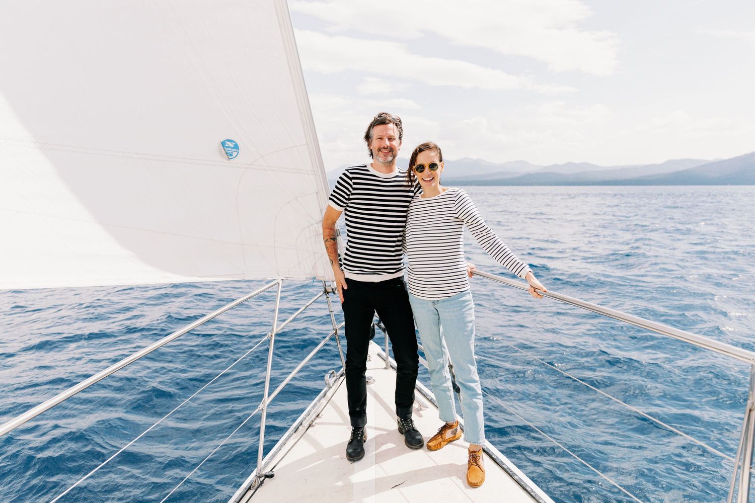 Two people in striped shirts stand together on the bow of a sailboat on blue ocean waters.