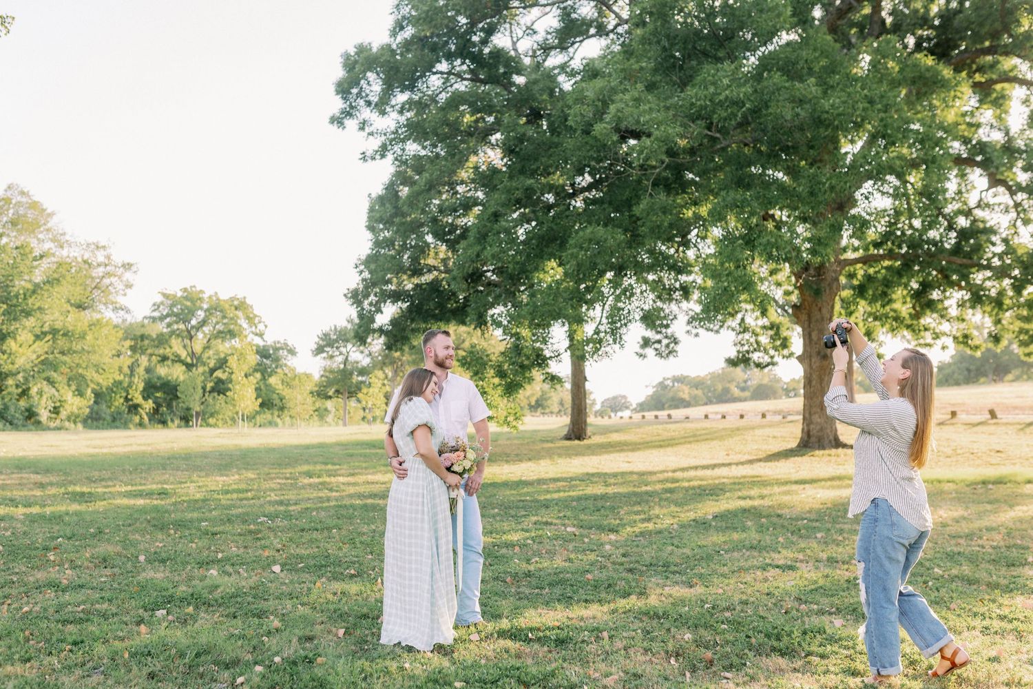 Two people stand in a sunny park setting with green grass and tall oak trees on a summer afternoon.