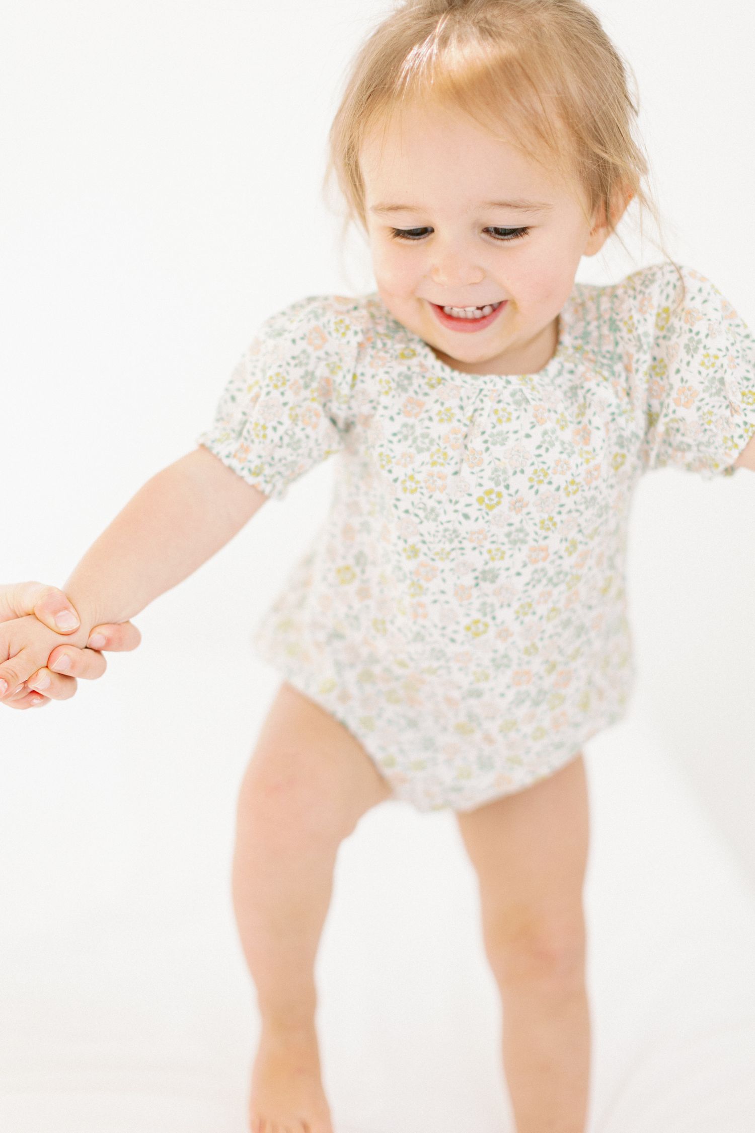 Happy toddler wearing floral print onesie reaches for something off-camera against white background.