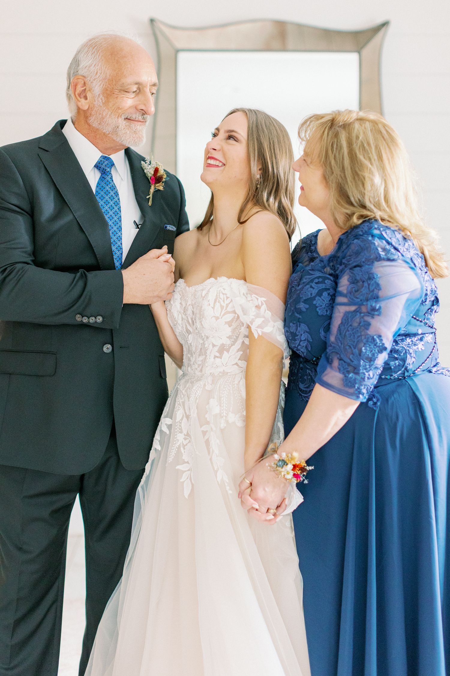 Three people share a joyful moment at a wedding, with the bride in white and others wearing blue and black formal attire.