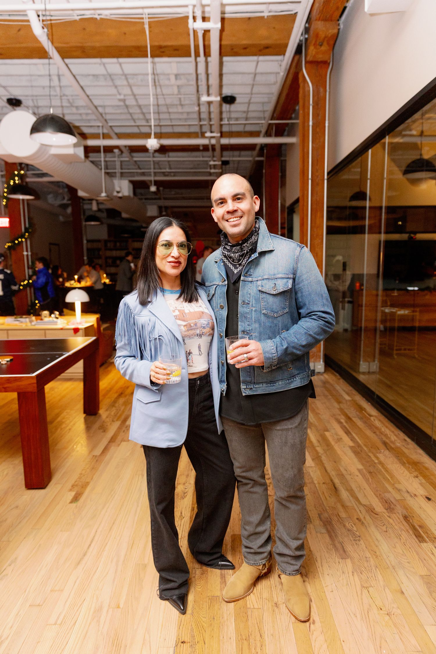 Two people pose together holding beverages in an industrial-style room with exposed pipes and wooden floors.