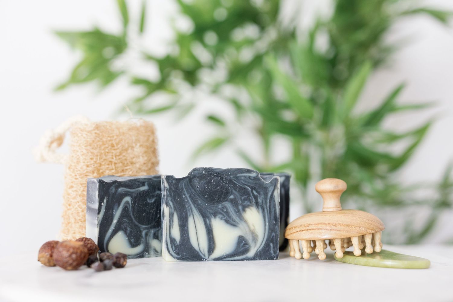 Handmade charcoal soap bar with natural spa accessories including loofah, wooden brush and green plants in background.