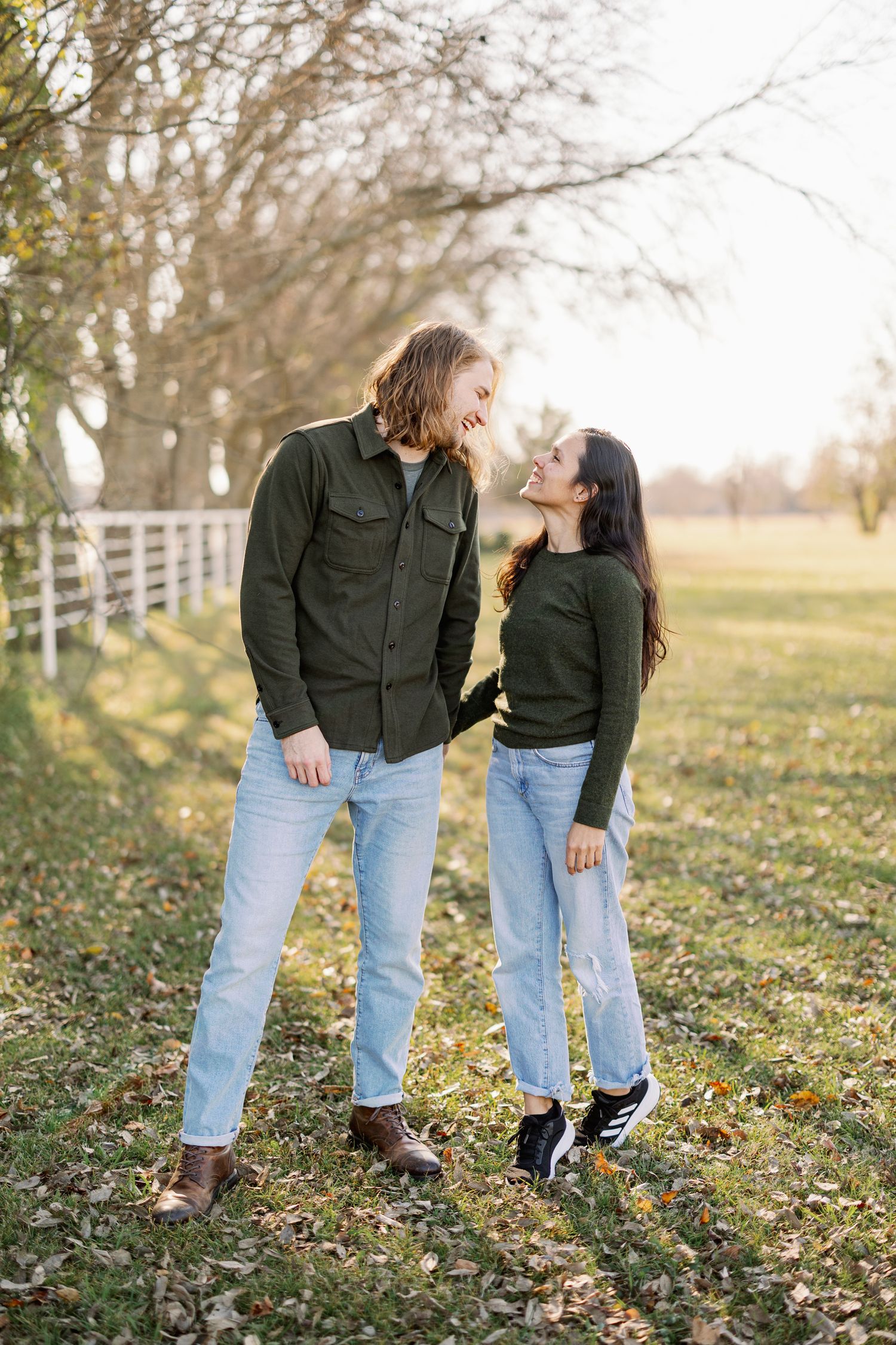 A couple in matching green shirts and jeans share a tender moment outdoors on an autumn day by a white fence.