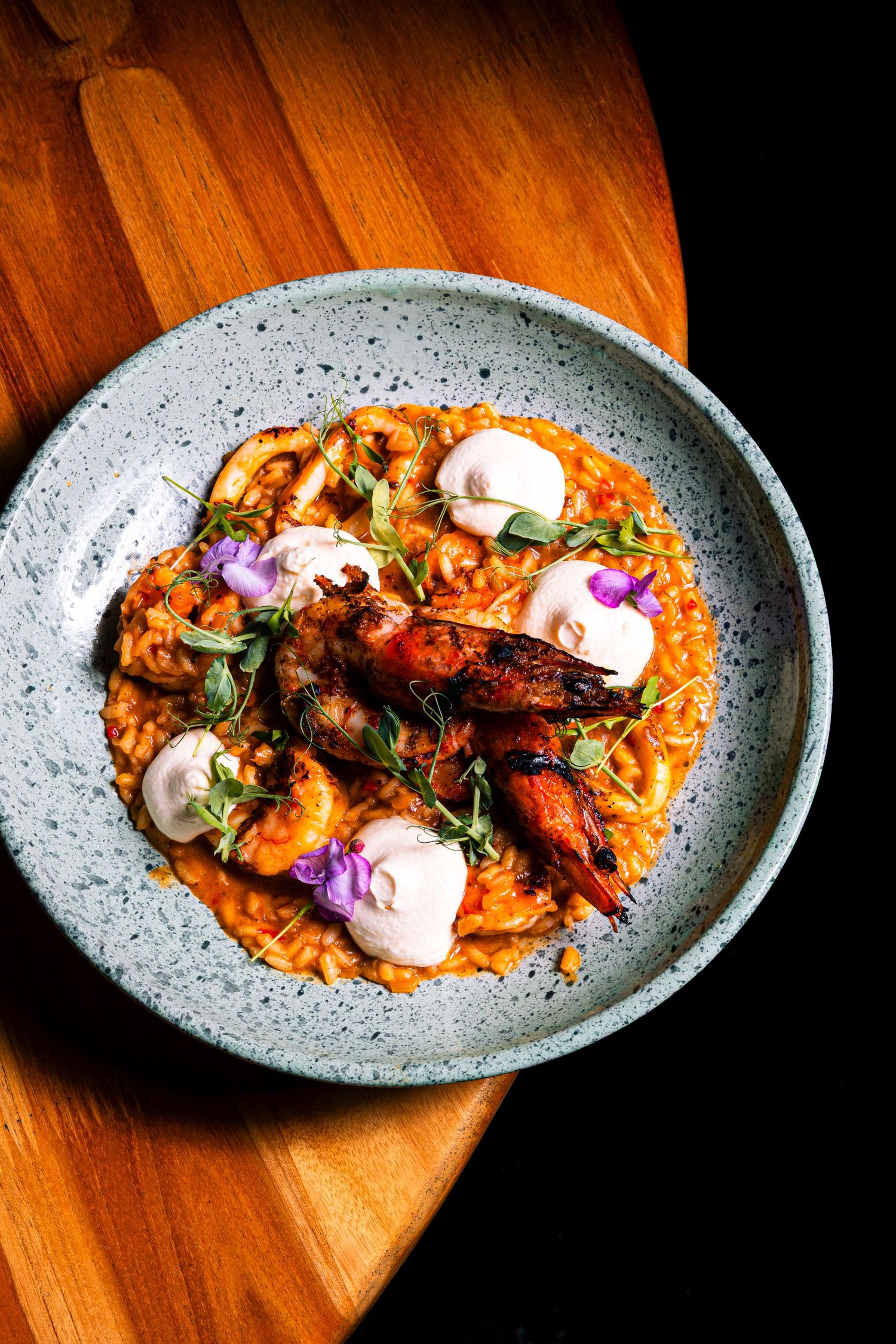Grilled octopus served on a speckled gray plate with creamy sauce and purple edible flowers on a wooden surface.