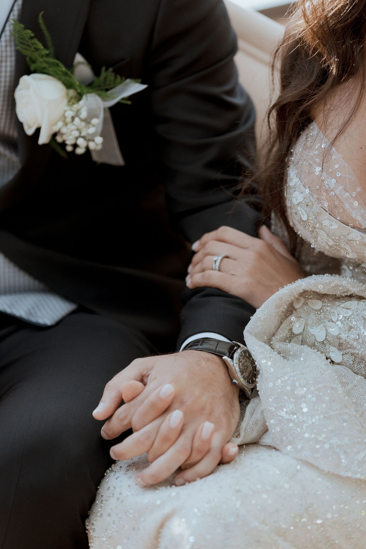 Close-up of bride and groom holding hands, showing wedding ring, watch, and floral boutonniere.