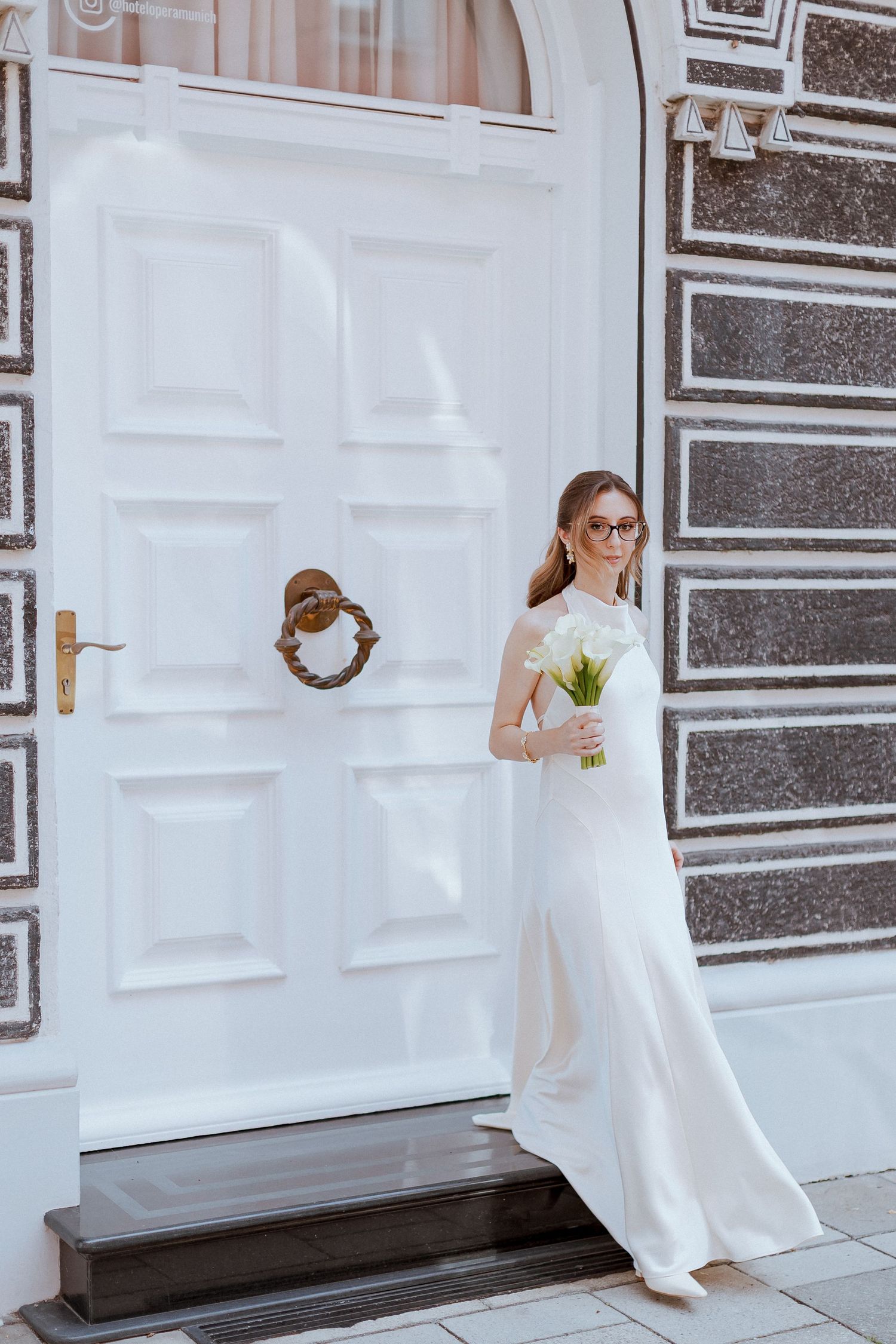 Elegant bride in white dress holding calla lily bouquet standing before classic white door.