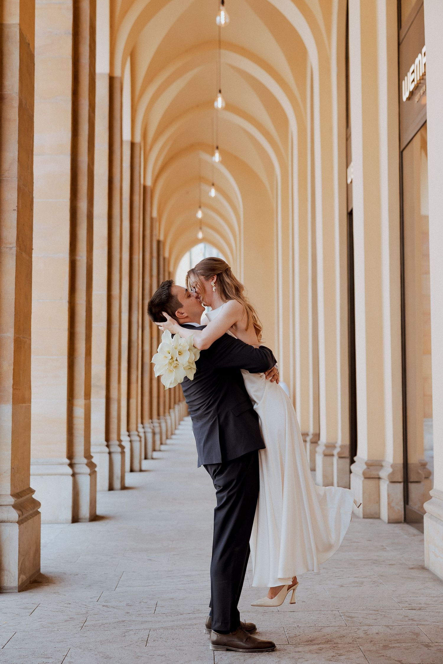 Newlywed couple shares romantic kiss in elegant arched hallway with repeating columns and dramatic lighting.