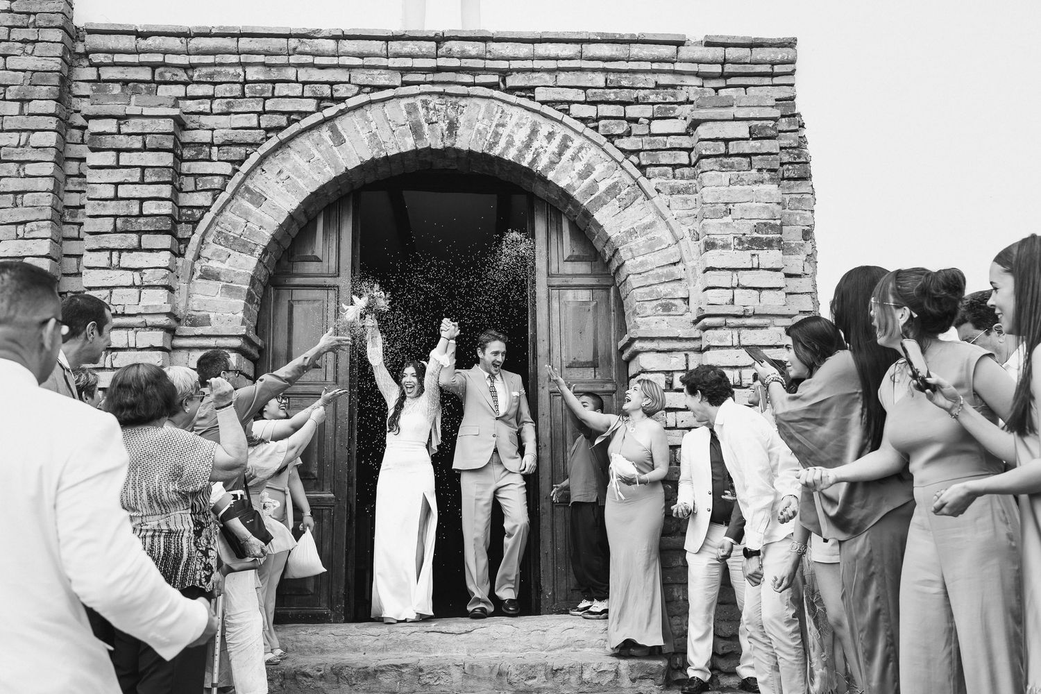 A black and white photo of a wedding couple exiting a stone church as guests throw rice in celebration.