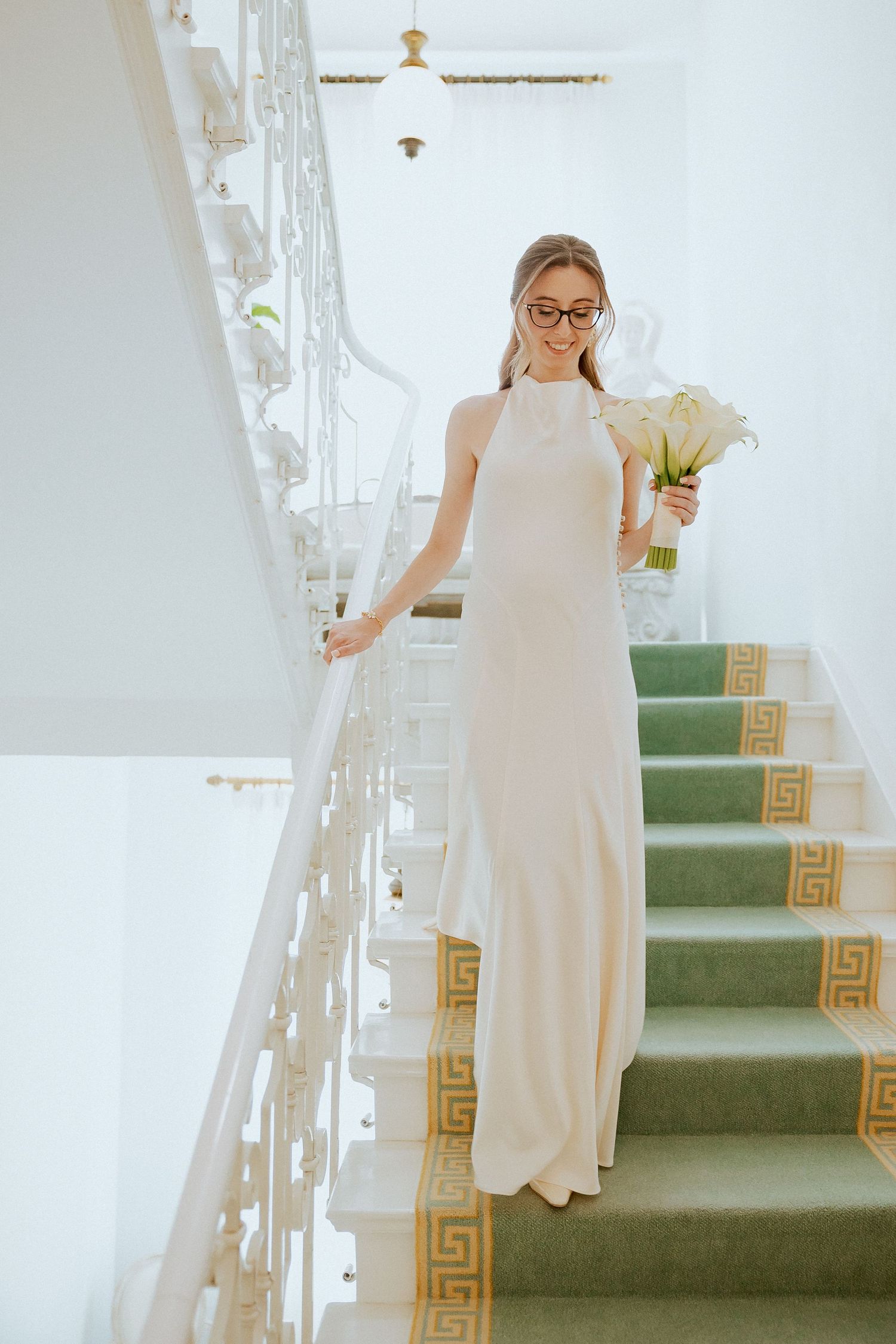Elegant bride in white halter gown holding bouquet descends ornate staircase with green carpet runner.
