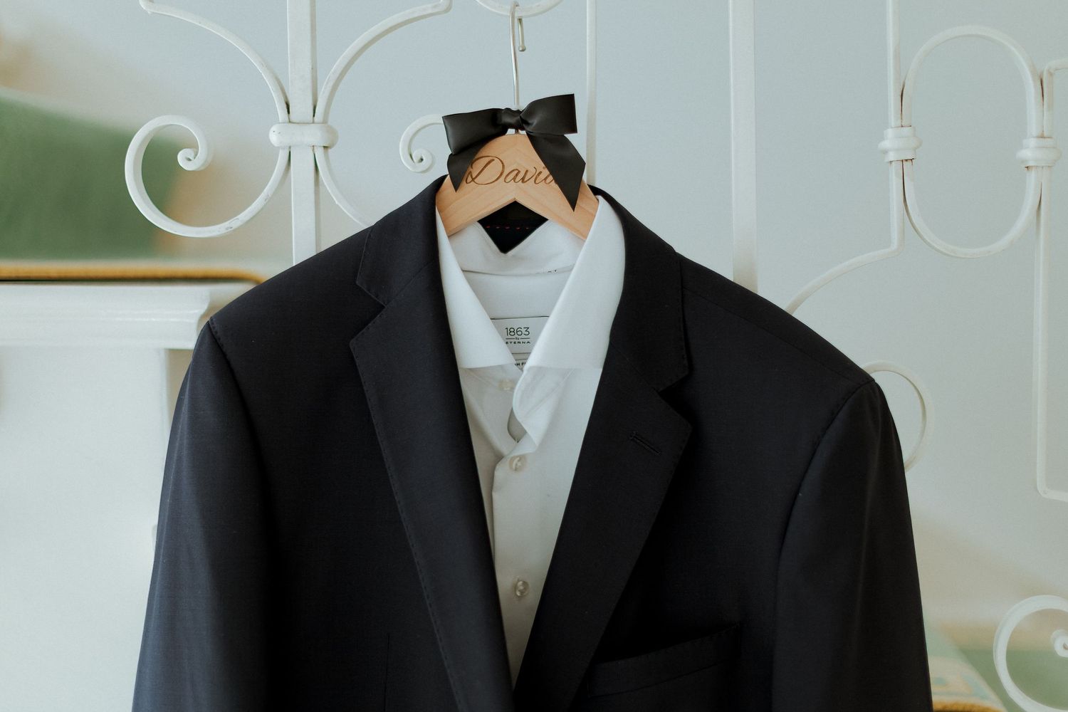 Black tuxedo jacket with white vest on a personalized wooden wedding hanger displayed on a decorative white frame.