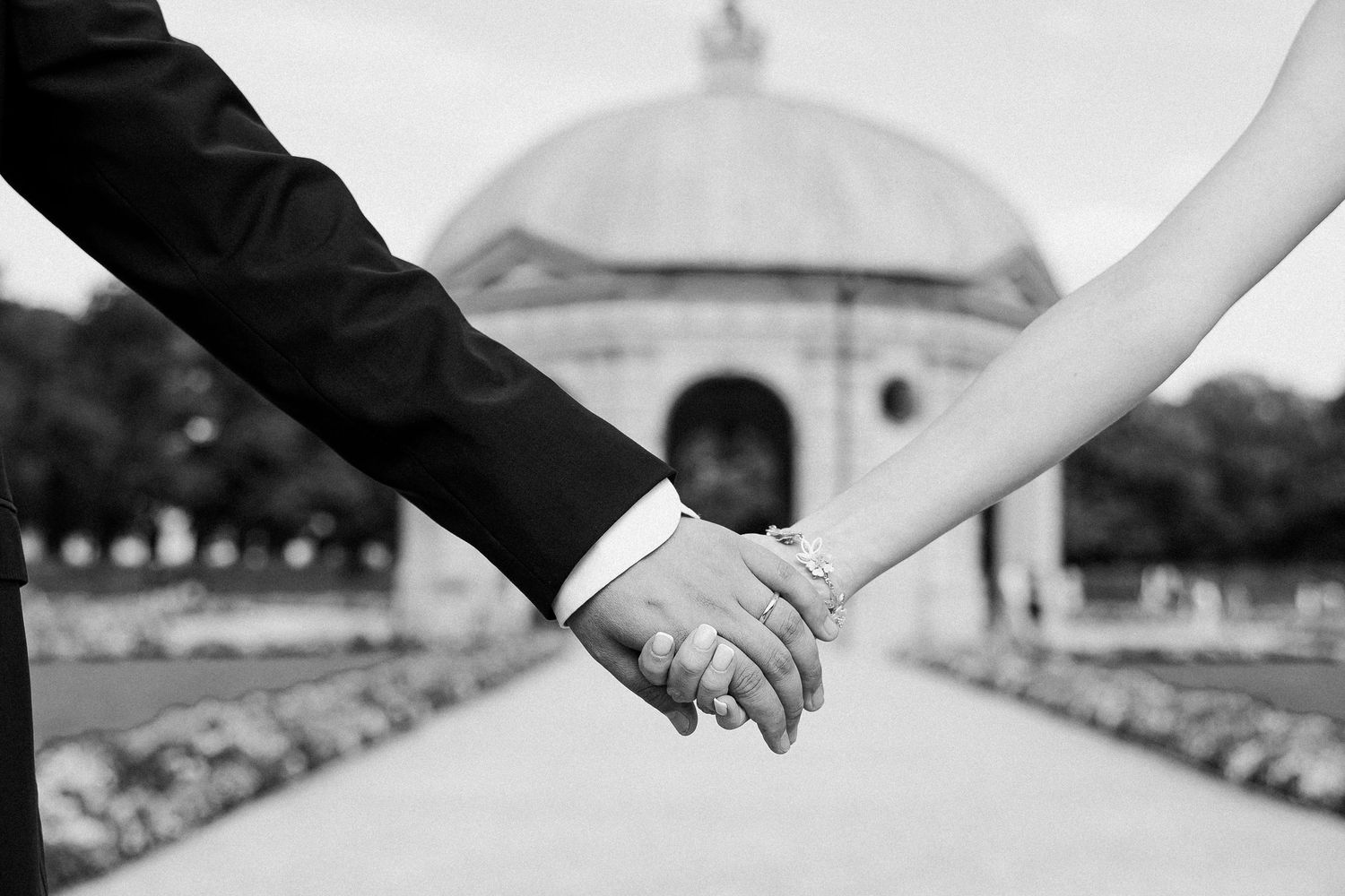 Two figures in wedding attire hold hands in close-up black and white shot of their hands together.