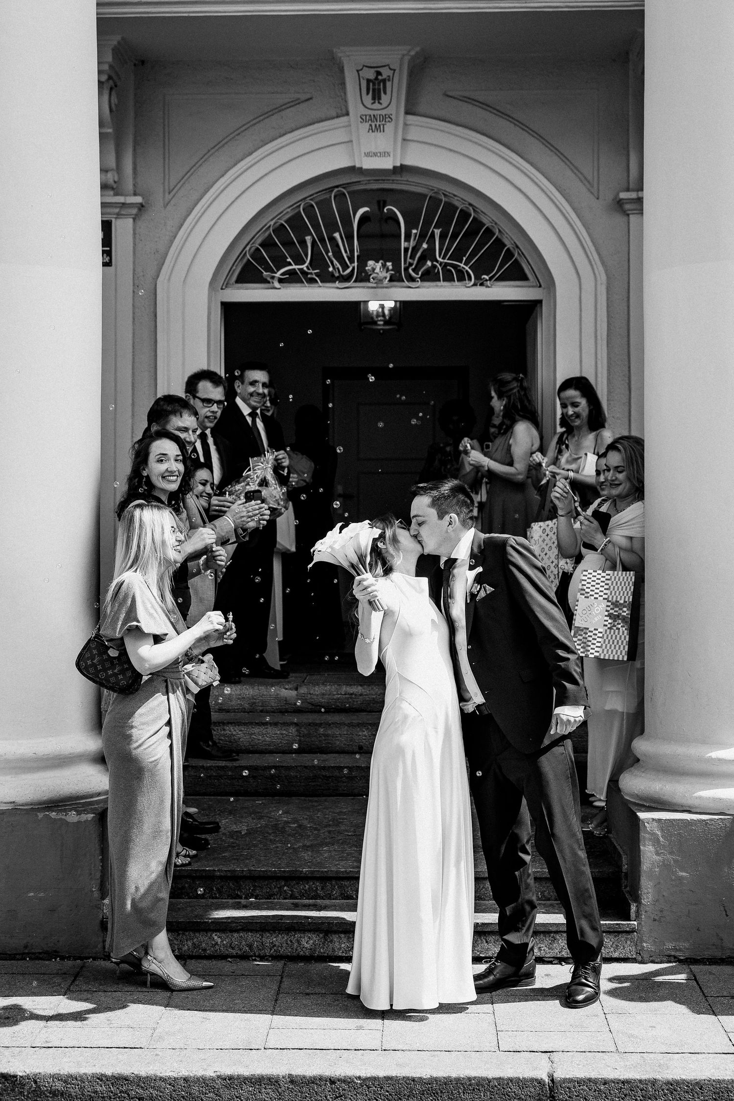 Newlyweds share a kiss on church steps surrounded by cheering guests in elegant black and white wedding photo.