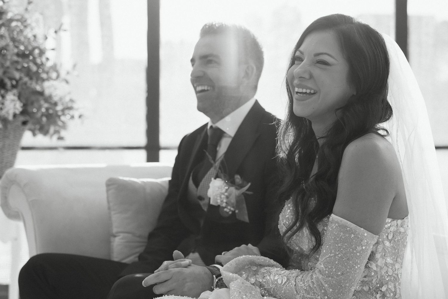 Bride and groom laughing joyfully together on a white sofa during their wedding ceremony in black and white photo.