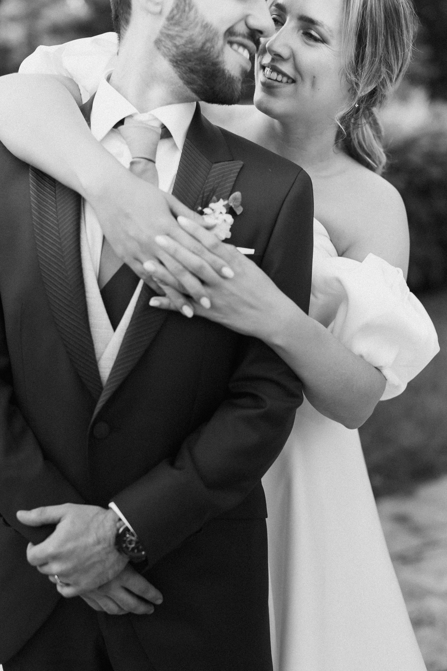 Black and white photo of smiling bride in white dress embracing groom in suit with boutonniere at wedding.