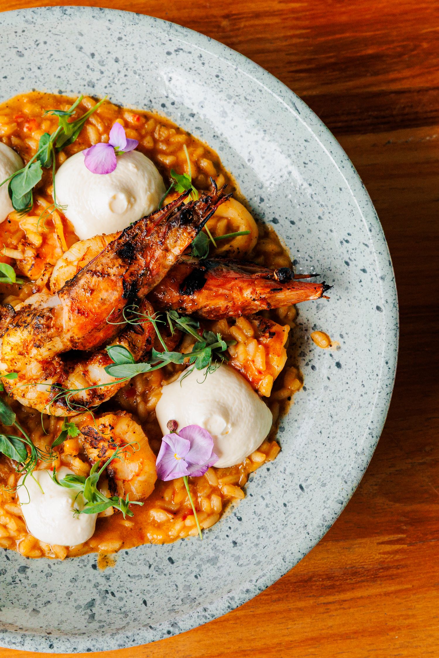 Grilled shrimp served on creamy risotto with dollops of white sauce and purple edible flowers on a speckled gray plate.