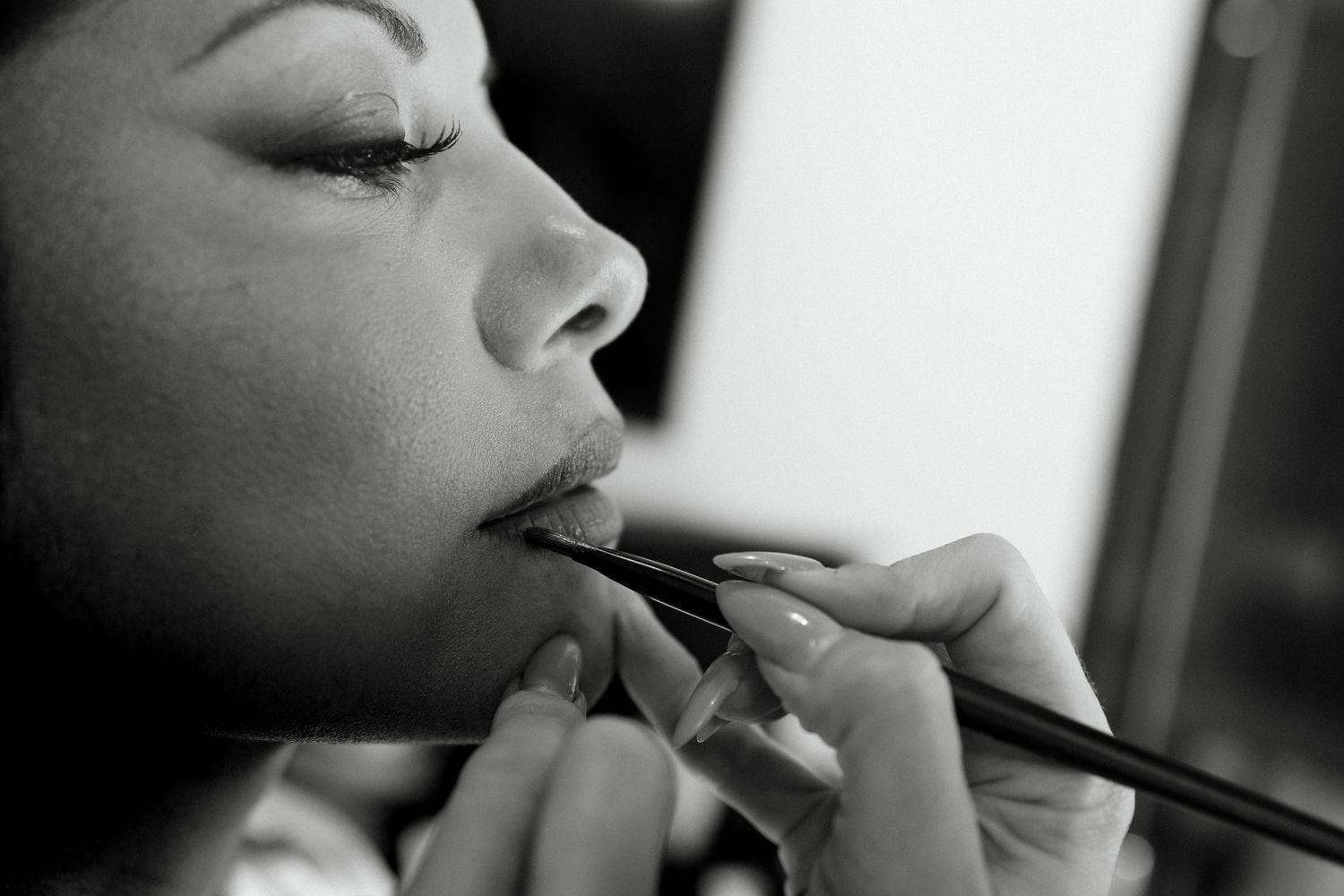 Black and white close-up of a makeup artist applying lipstick with a brush to a woman's lips.