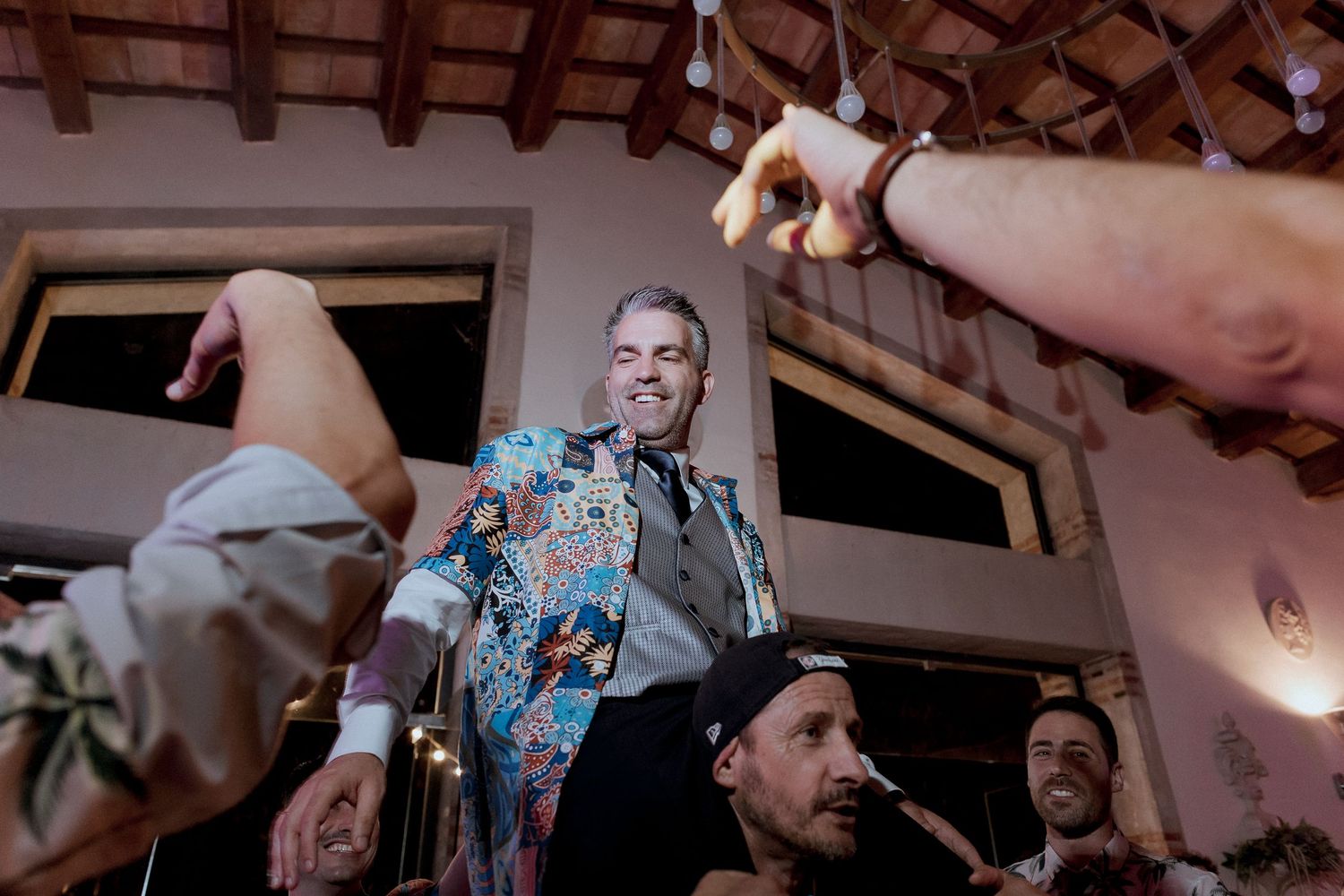 Groom in colorful floral blazer celebrated by guests at lively indoor wedding reception party.