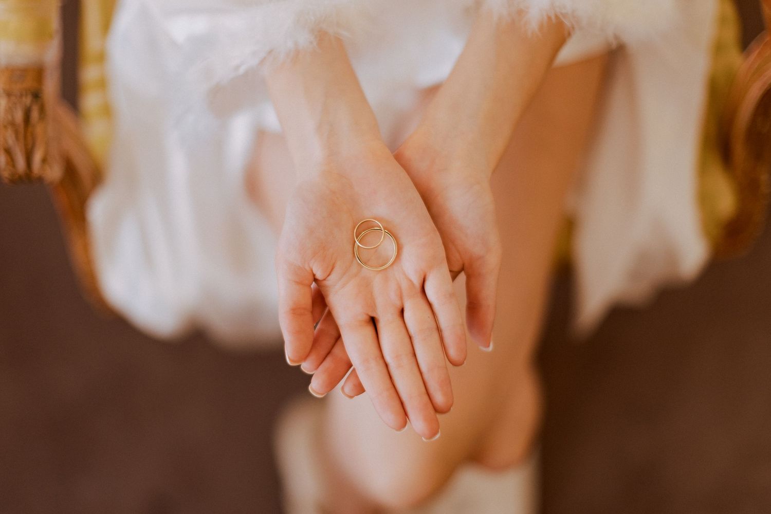 Close-up of hands holding a delicate gold ring, wearing a white dress with soft bokeh background.