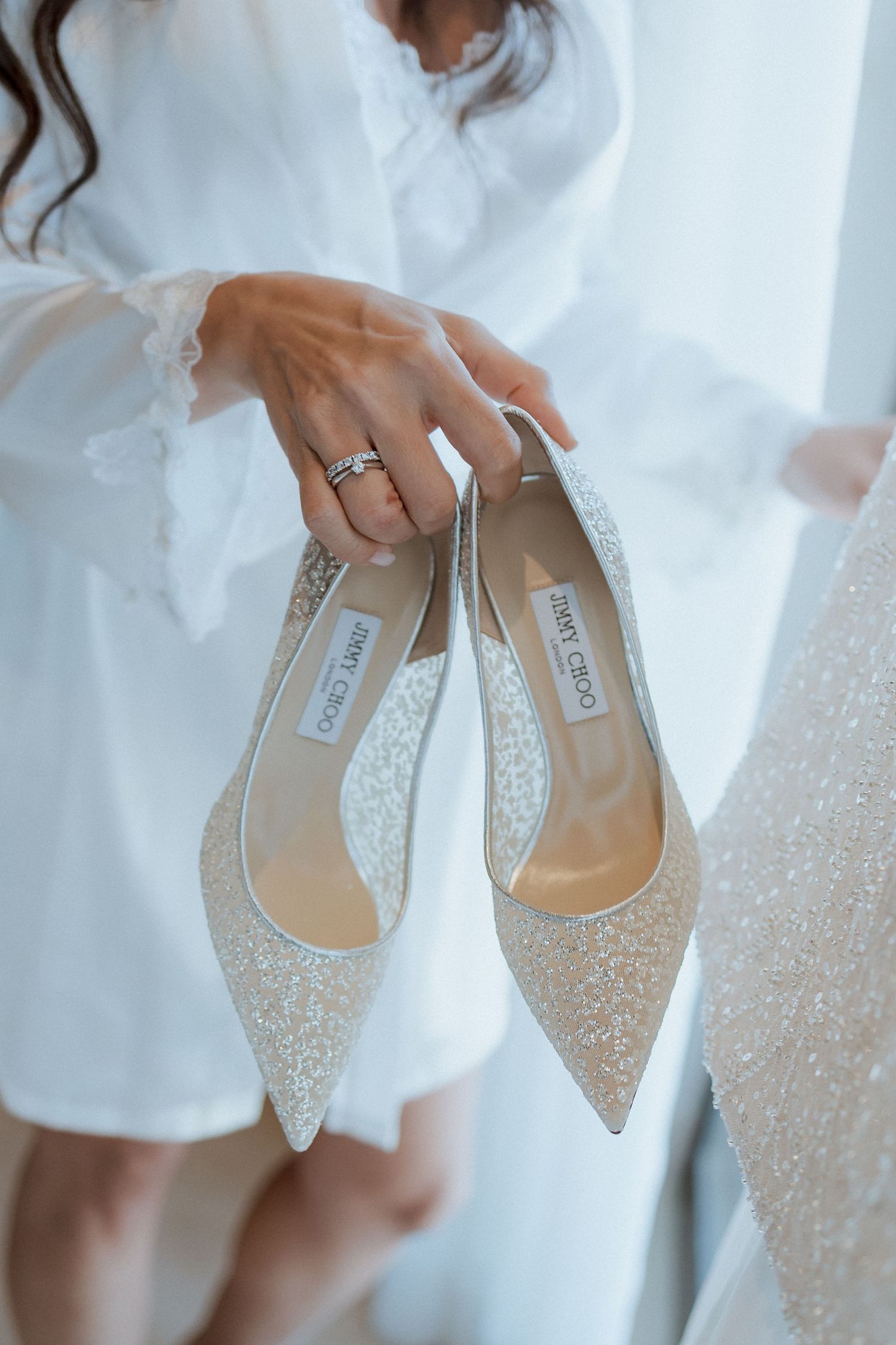 Bride in white robe holding sparkling Jimmy Choo pointed-toe wedding heels with engagement ring visible.