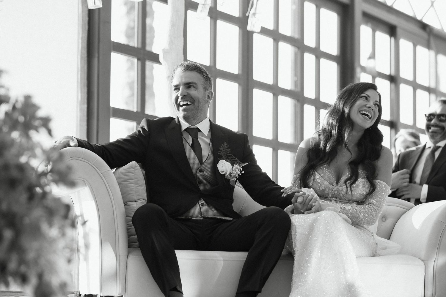 Black and white photo of a laughing bride and groom sitting together on a white sofa during their wedding ceremony.