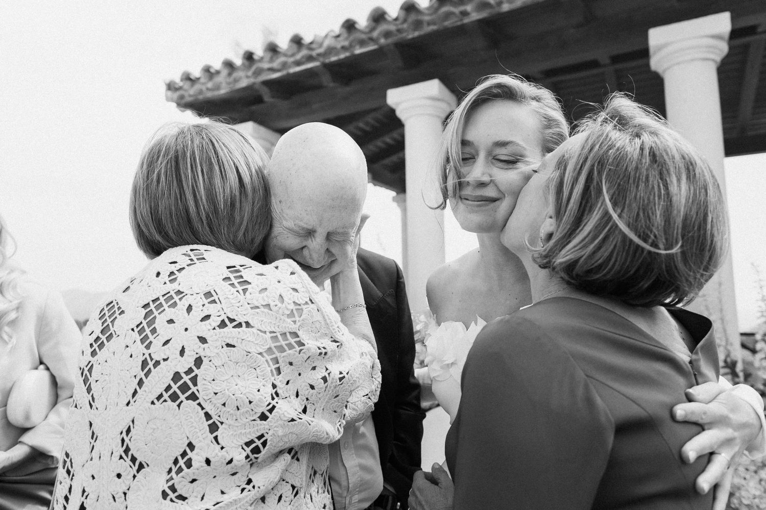Black and white wedding photo of bride receiving emotional hugs from family members outdoors near classic columns.