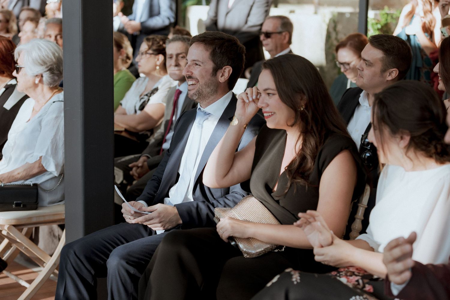 Guests laughing and smiling while seated at an outdoor wedding ceremony.