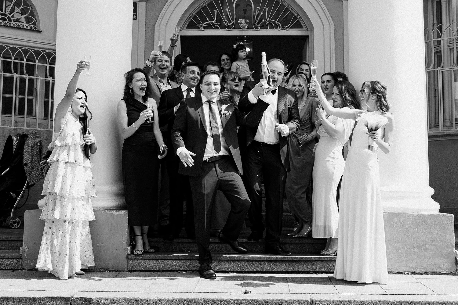Groom joyfully exits church steps surrounded by cheering wedding guests in elegant attire at a classic venue.