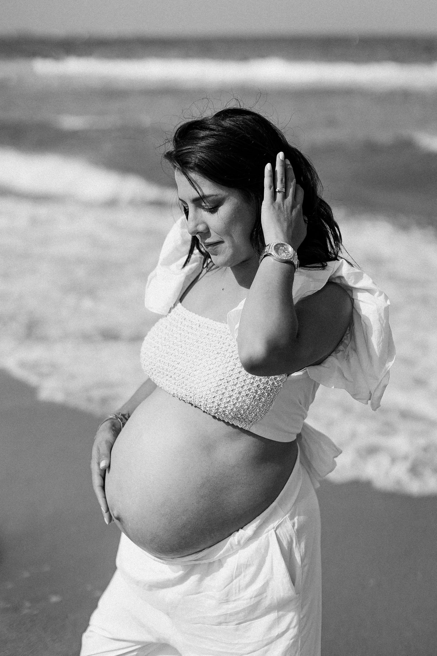 Black and white portrait of an expectant mother in a white dress at the beach with ocean waves in the background.