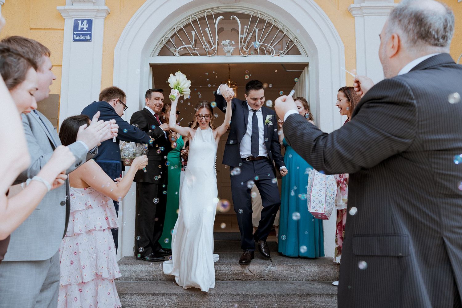 Joyful newlyweds exit a church ceremony as guests celebrate with confetti and bubbles in a sunny outdoor wedding.