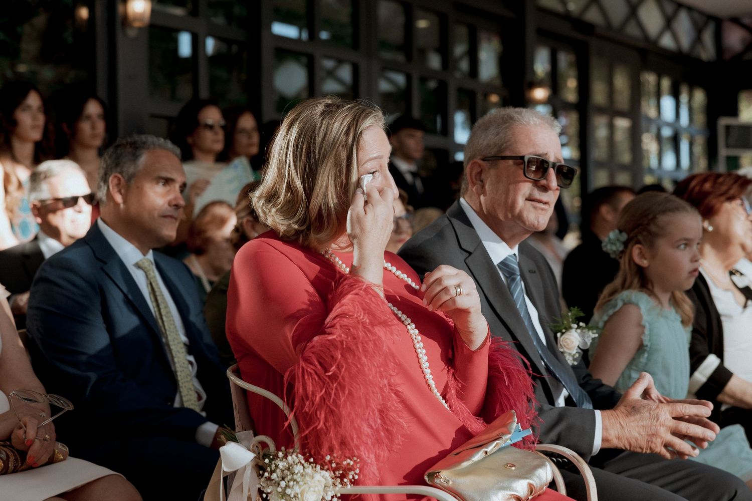 Emotional wedding guests laughing and crying during ceremony, woman in red dress wiping tears, elderly man in sunglasses smiling.