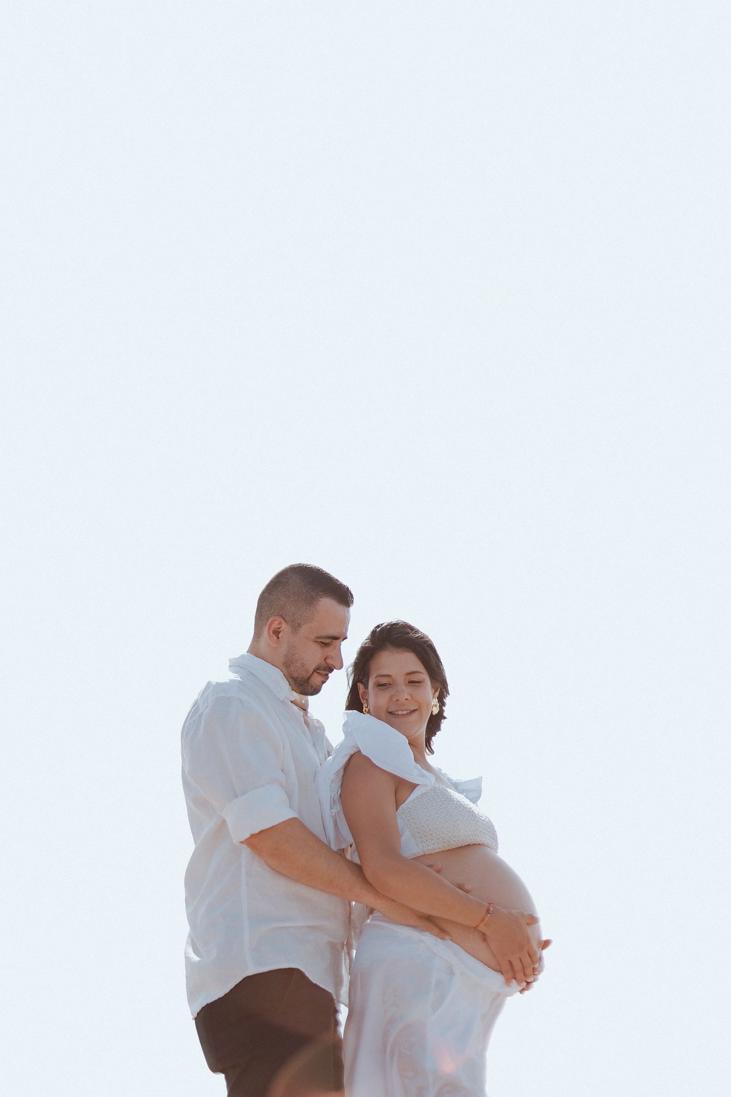 A couple dressed in white embraces outdoors during a maternity photoshoot against a bright sky background.