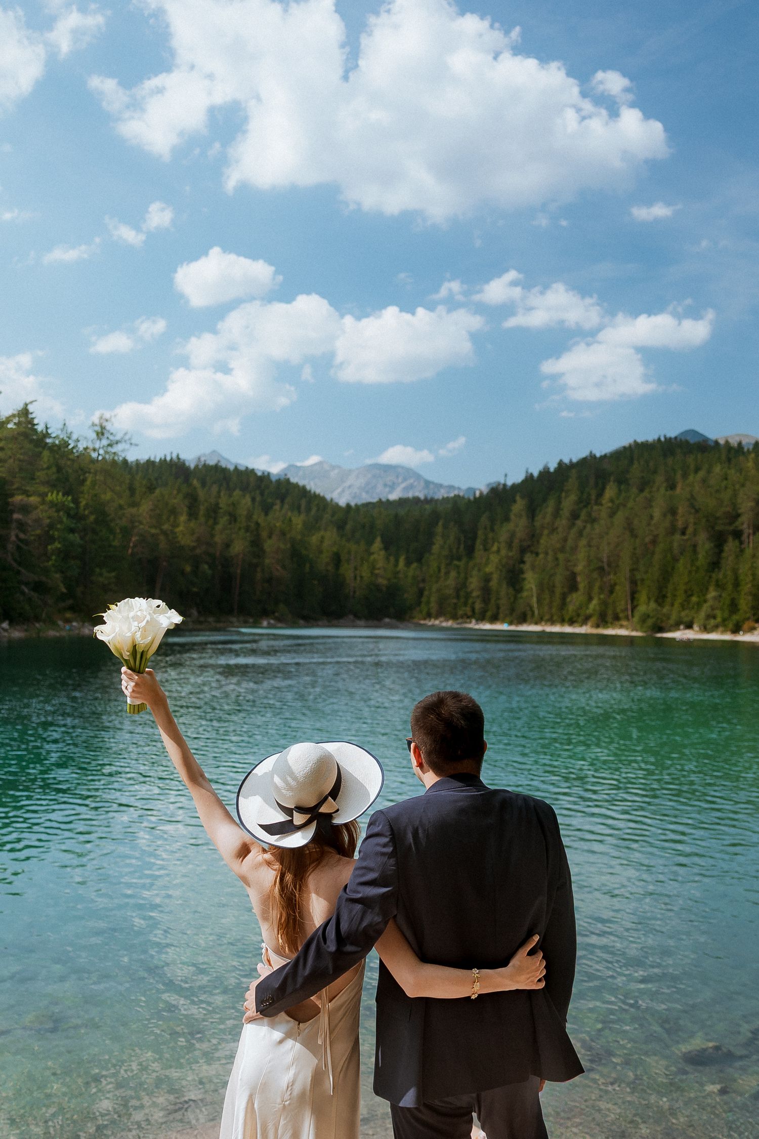 A couple in wedding attire sits by a serene mountain lake surrounded by evergreen forest on a sunny summer day.