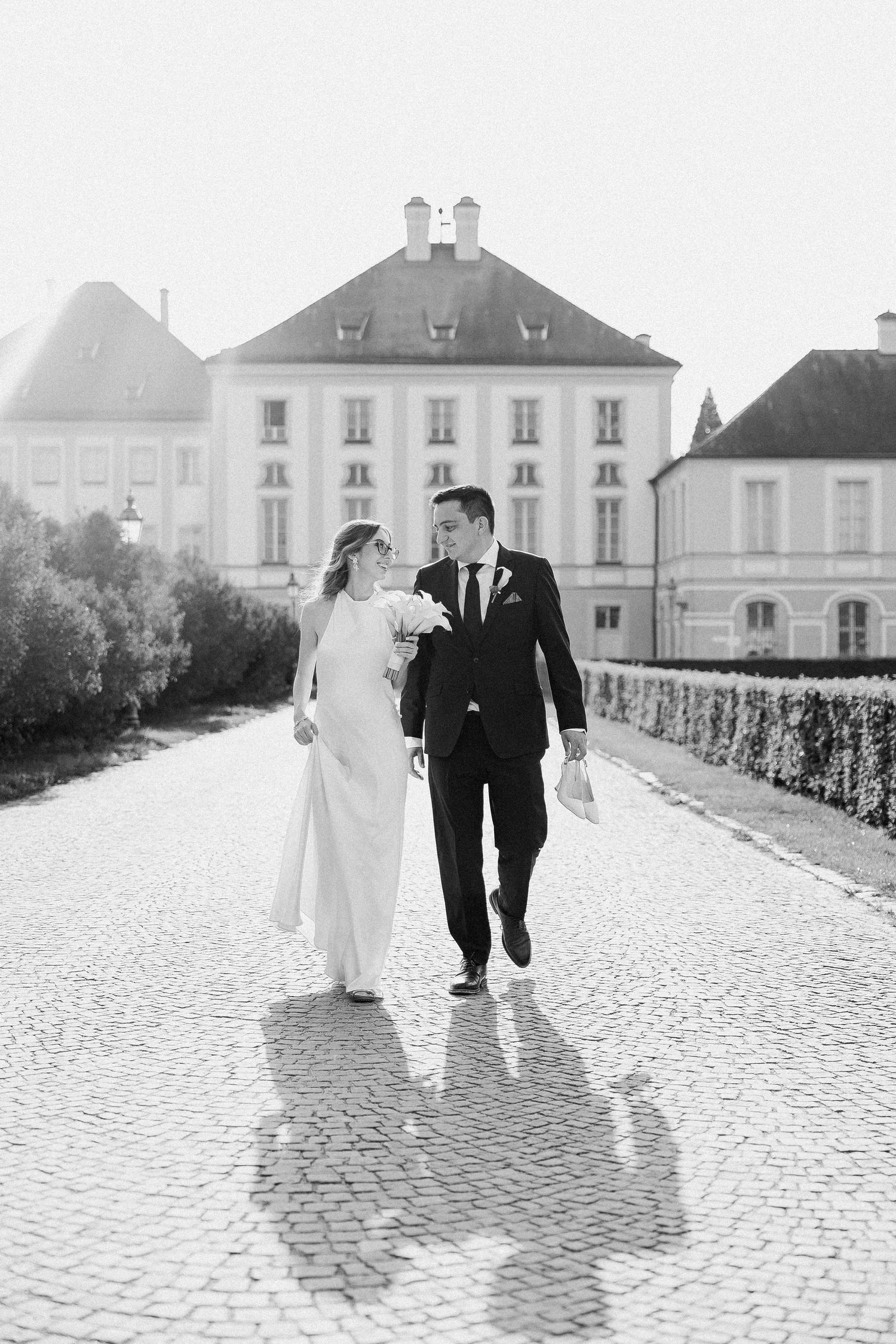 Bride and groom walk hand in hand on cobblestone path before a grand European manor in black and white.