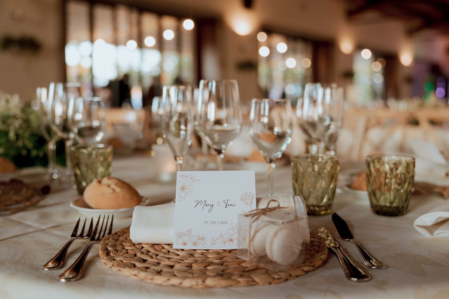 Elegant wedding reception table setting with wicker charger, glassware, greenery, and personalized place card.