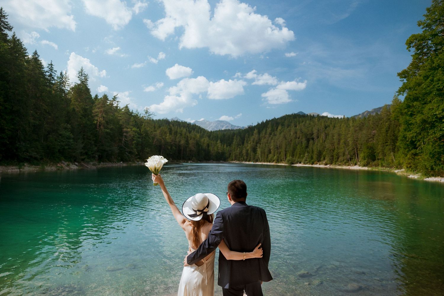 Newlywed couple standing by a turquoise mountain lake surrounded by pine forests on their wedding day.