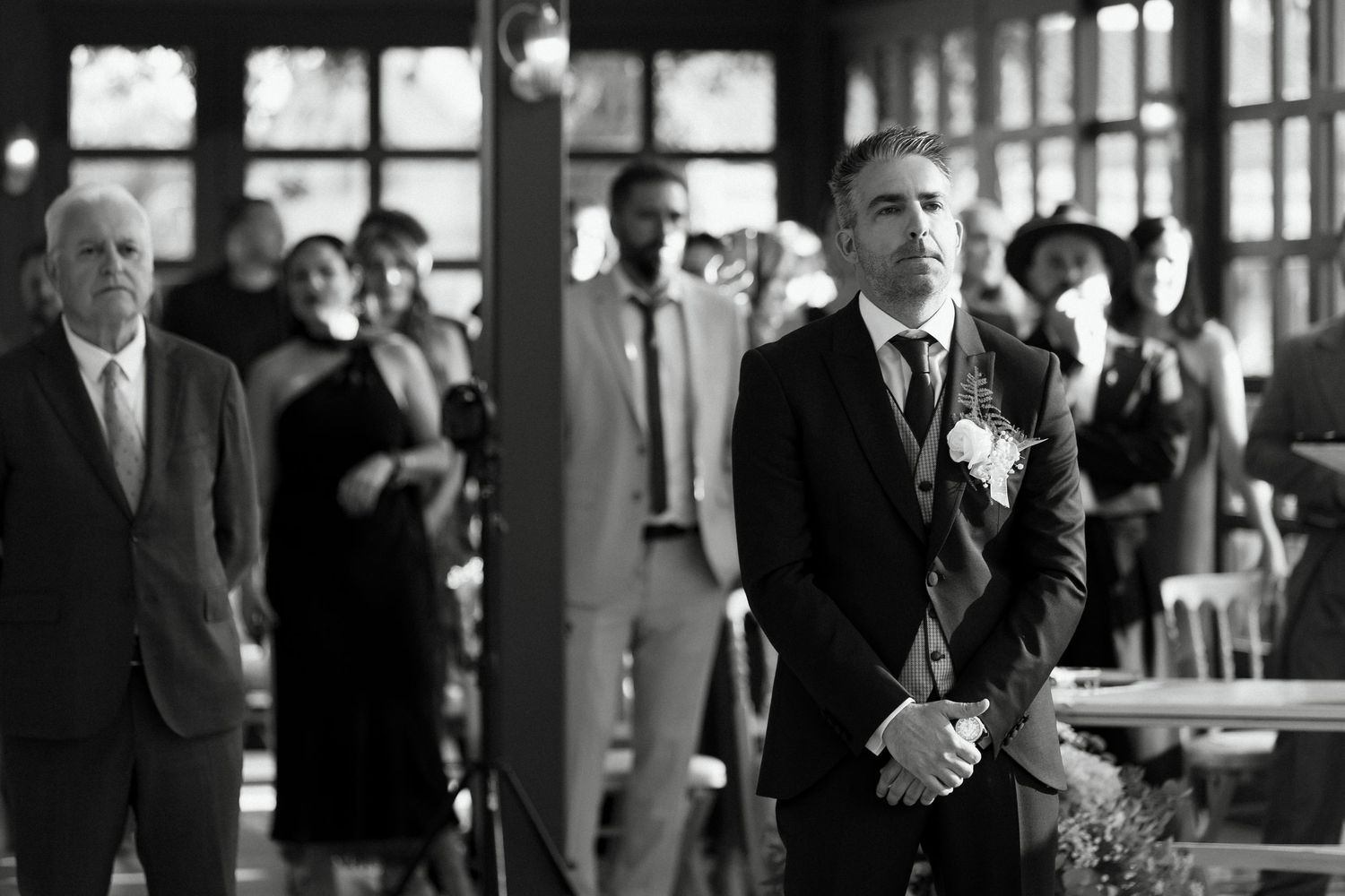 Groom in black suit with boutonniere waits at altar during wedding ceremony in elegant venue.