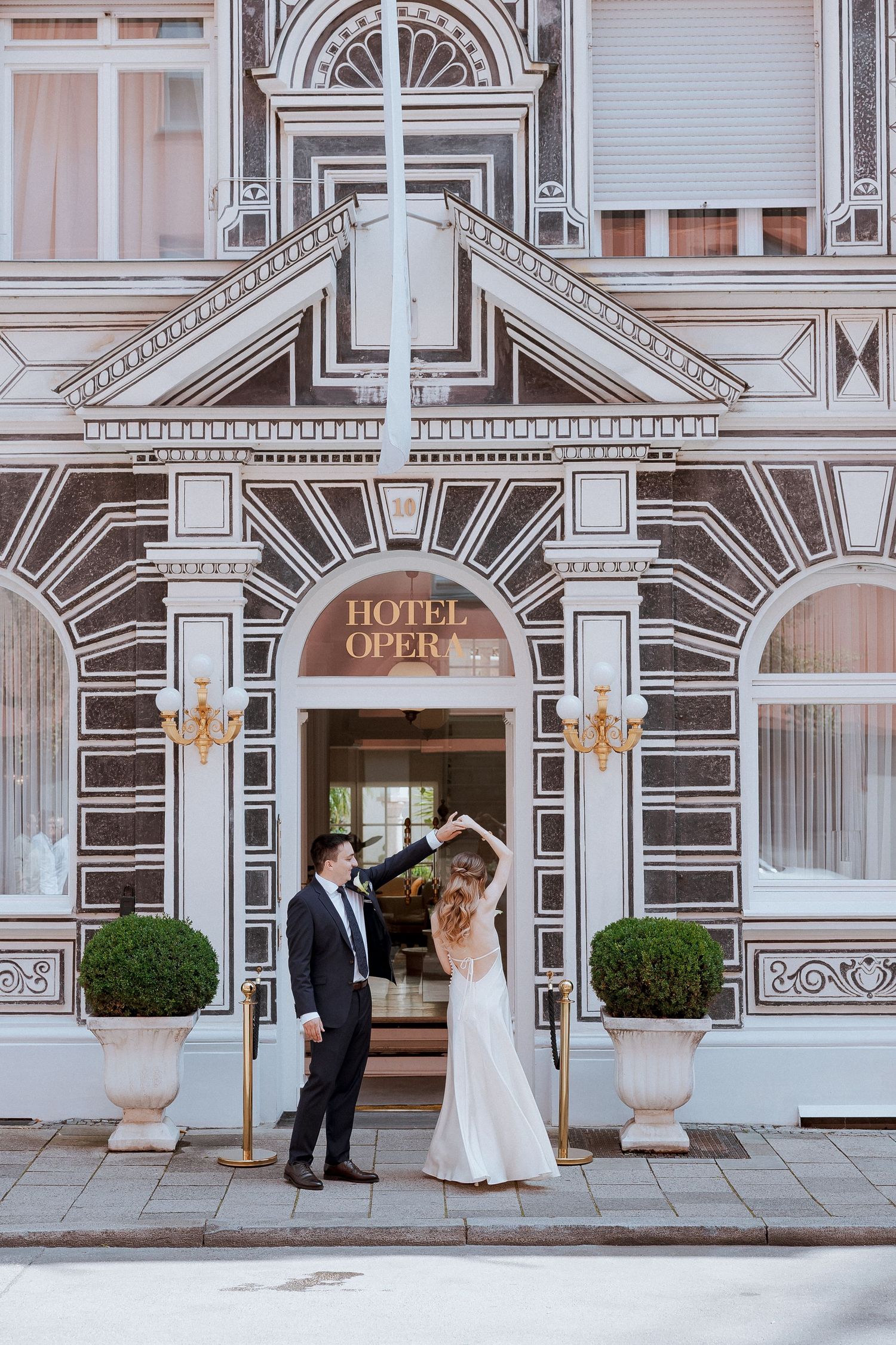 Bride and groom dancing in front of Hotel Opera, bride in white gown, groom in black suit, elegant facade.