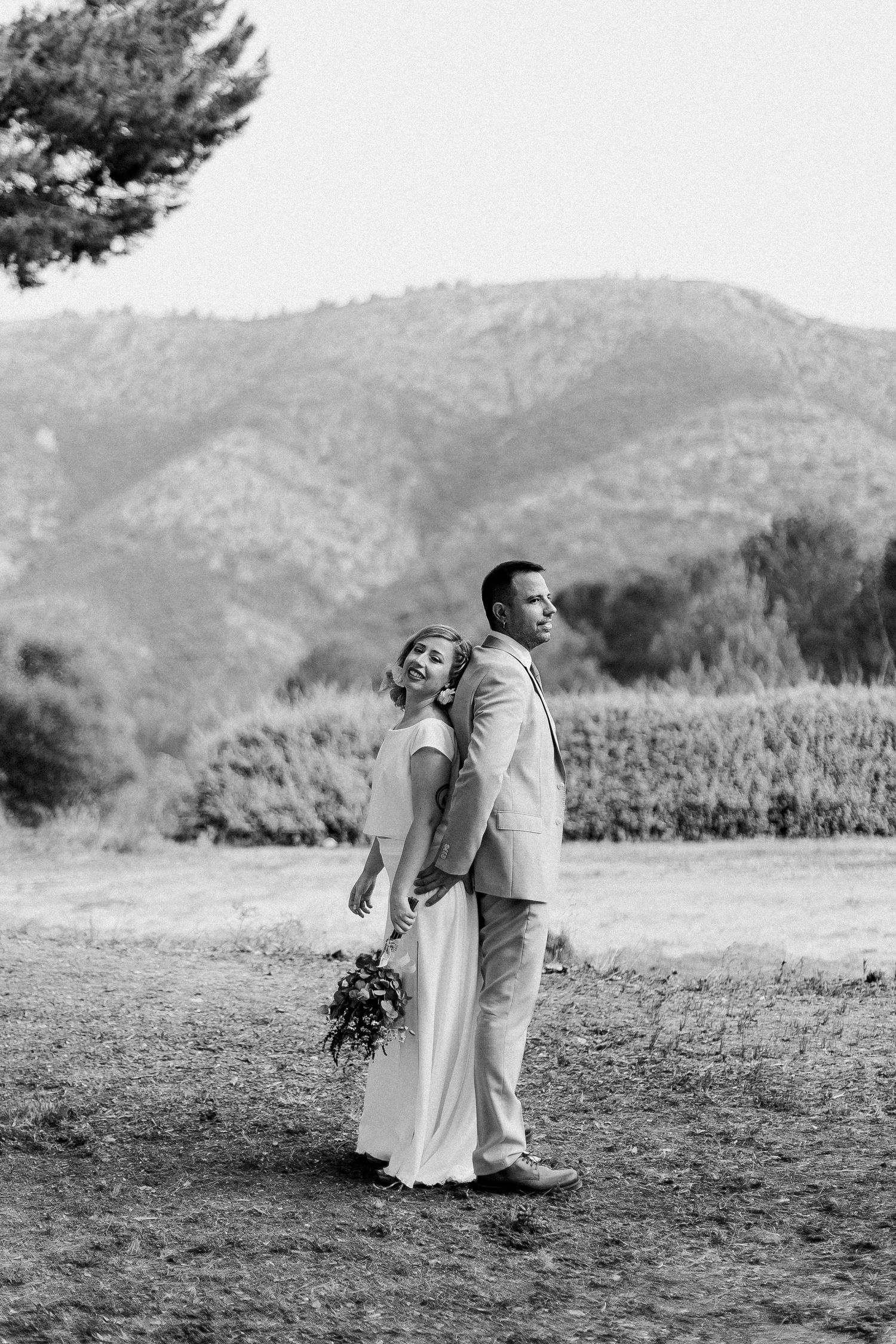 A couple poses back-to-back outdoors in formal wedding attire with mountains and trees in the background.