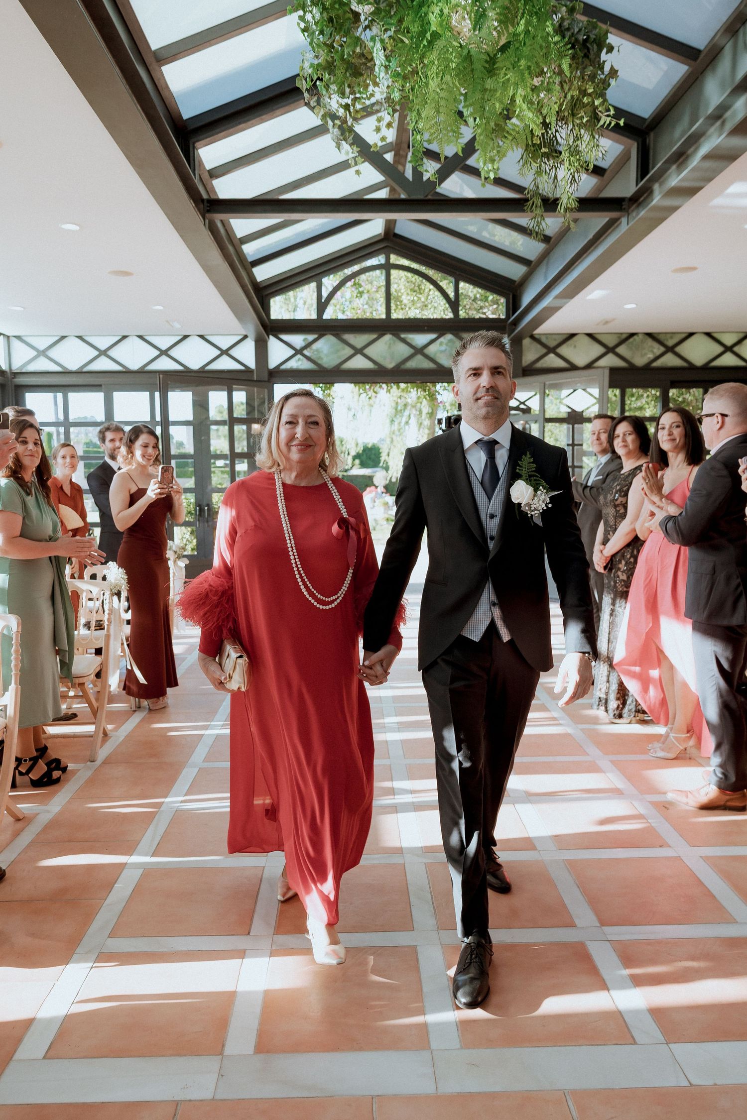 Groom in black suit walking down the aisle with woman in red dress in a glass-roofed venue.