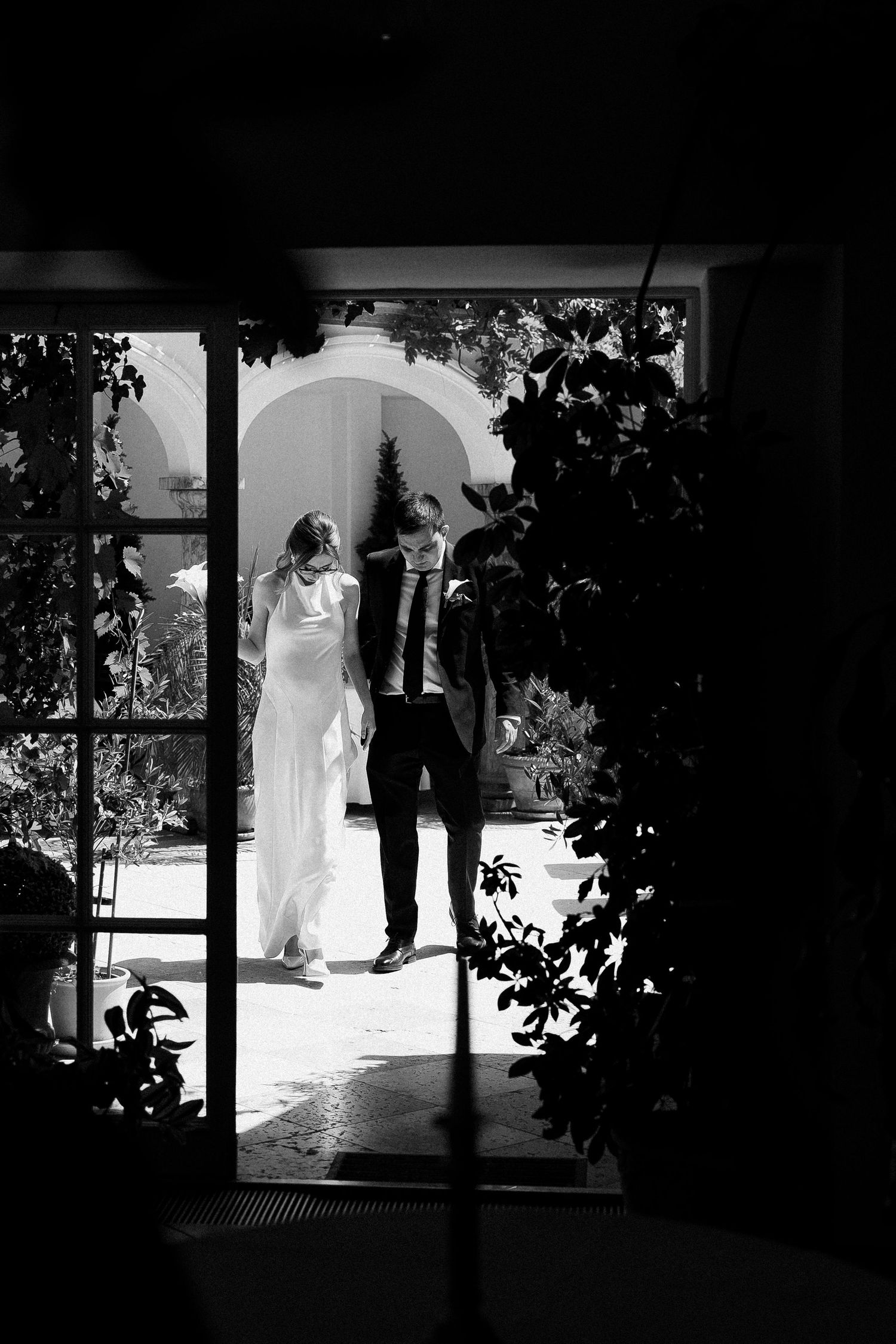Black and white photo of a bride and groom silhouetted in a sunlit doorway surrounded by lush greenery.