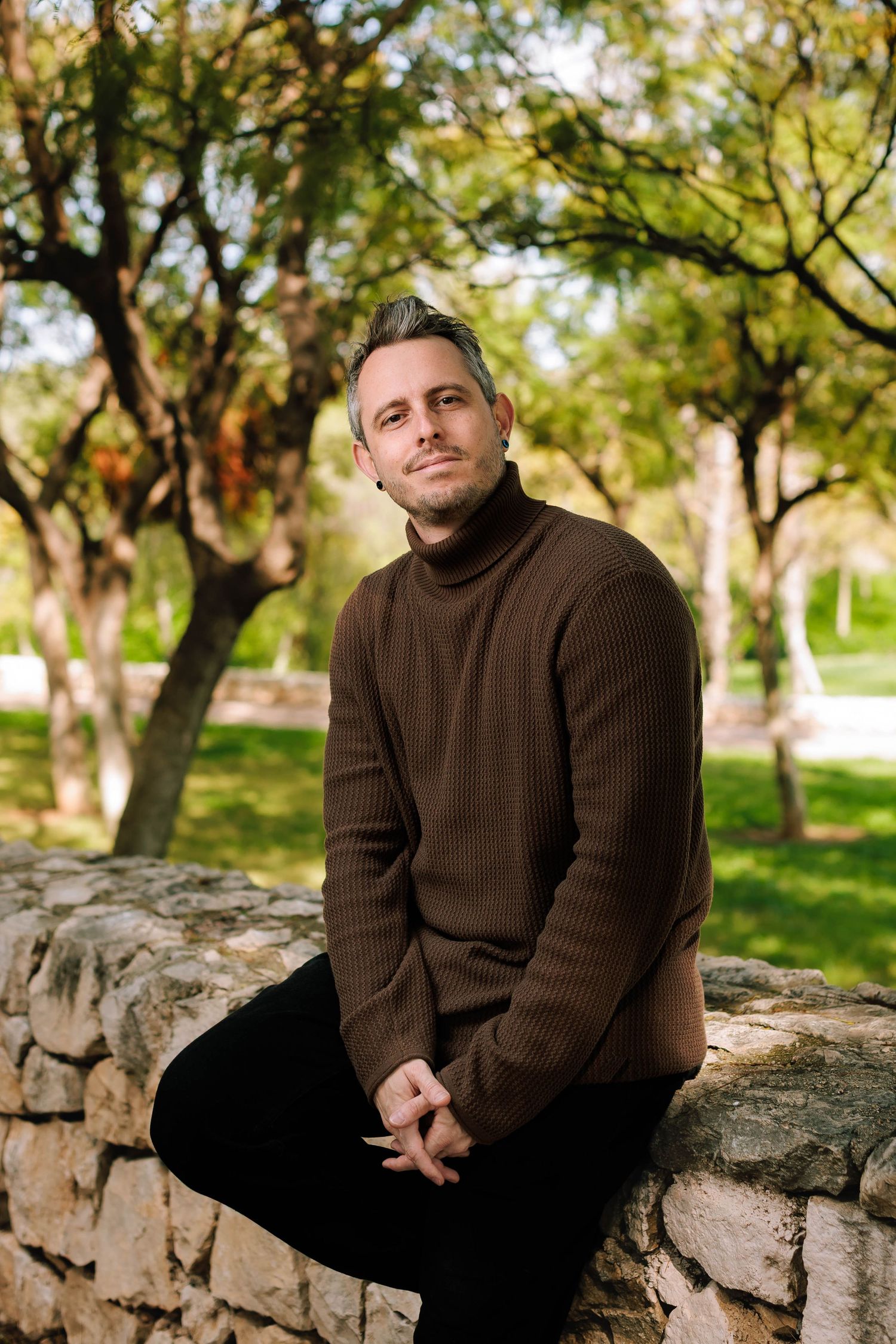 A person wearing a brown sweater sits on a stone wall in a park setting with autumn trees in the background.