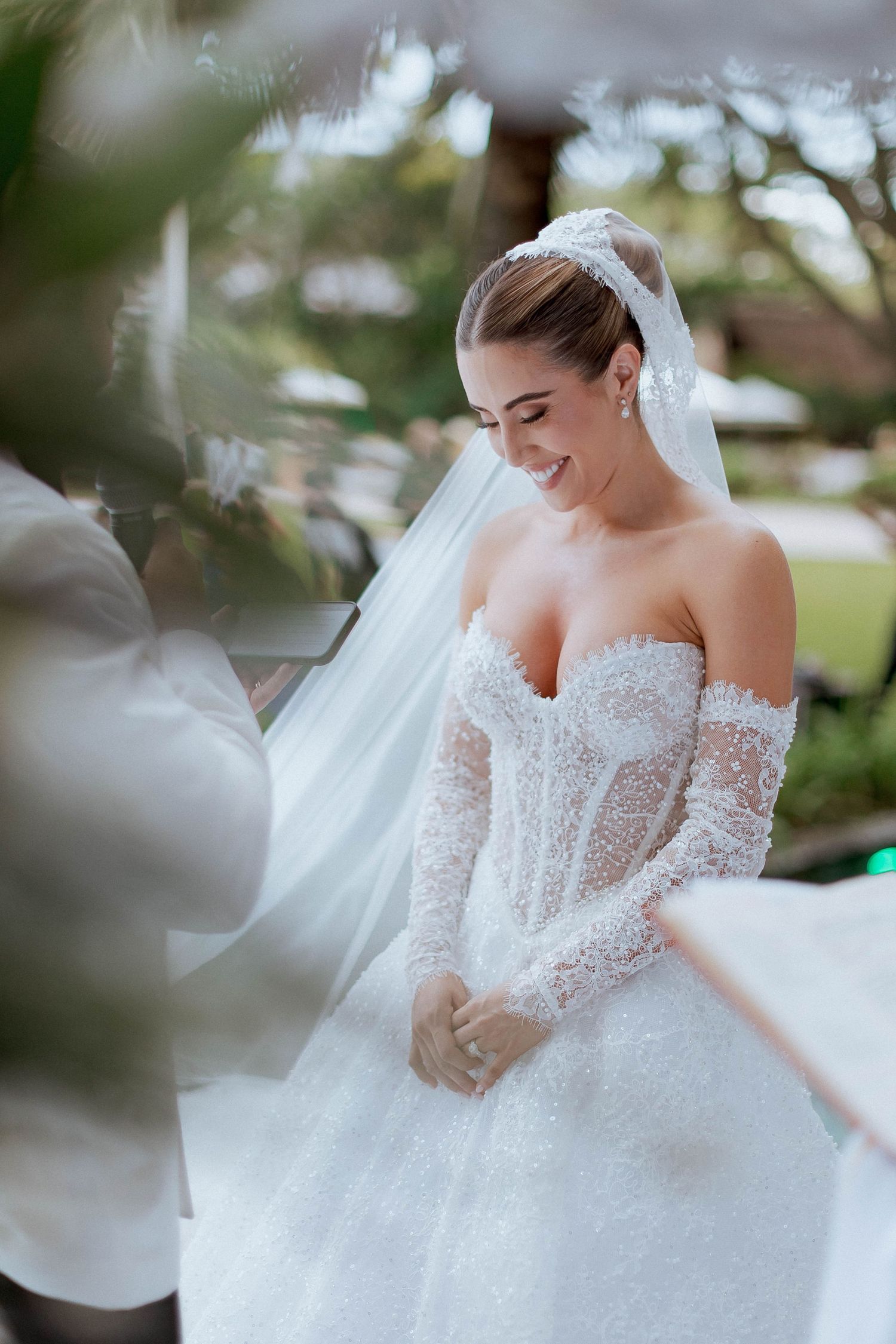 A bride in an elegant off-shoulder lace wedding gown with a flowing tulle skirt smiles during her outdoor photo session.